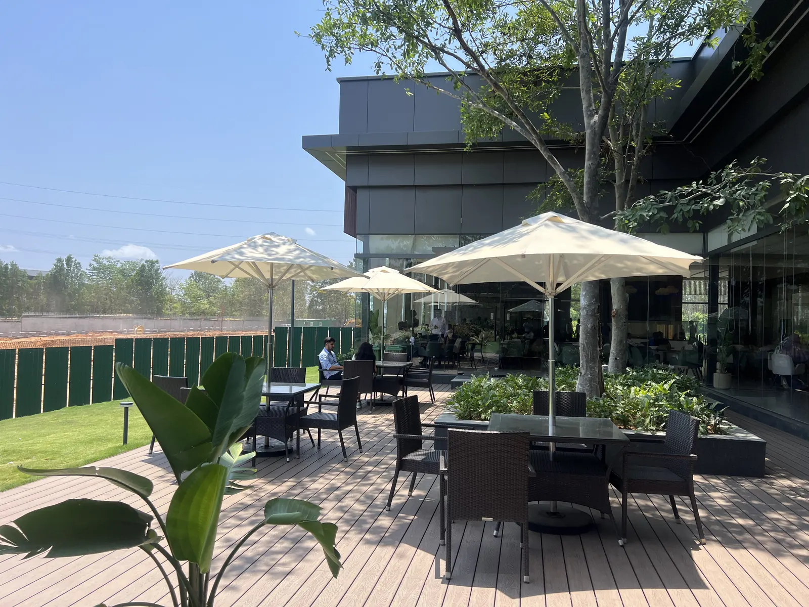 Featured Image of Godrej Vanantara Experience Centre outdoor cafe social zone with umbrella seating, timber decking and project-site landscaping &mdash; a preview of the planned clubhouse cafe